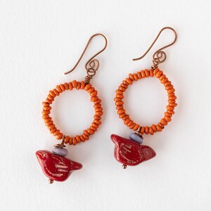 May include: A pair of earrings with copper-coloured ear wires and orange beaded hoops. Each earring has a red bird charm with gold detailing and a small purple bead. The earrings are set against a white background.