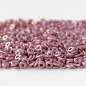 May include: A close-up shot of a collection of small, circular, mauve-coloured beads. Each bead has a hole in the centre and a slightly iridescent finish. The beads are scattered on a white surface, creating a textured effect.