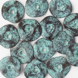 Może przedstawiać: Kolekcja turkusowych i brązowych koralików w kształcie dysków z centralnymi otworami. Koraliki mają zwietrzały, teksturowany wygląd, sugerujący starą lub antyczną estetykę. Paleta kolorów zdominowana jest przez odcienie turkusu i ciemnego brązu.