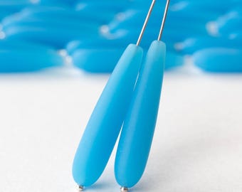 May include: Close-up of two light blue teardrop-shaped beads with silver-colored metal pins. The beads have a smooth, matte finish. Other similar beads are visible in the background, slightly out of focus.