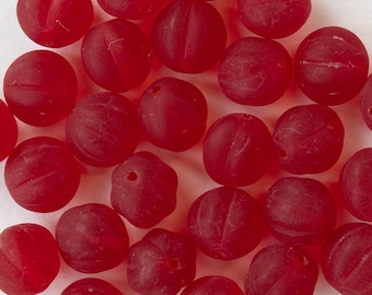 May include: A collection of round, red glass beads with a matte finish. Each bead has a grooved design and a small hole for stringing. The beads are scattered on a white surface, creating a visually appealing arrangement.