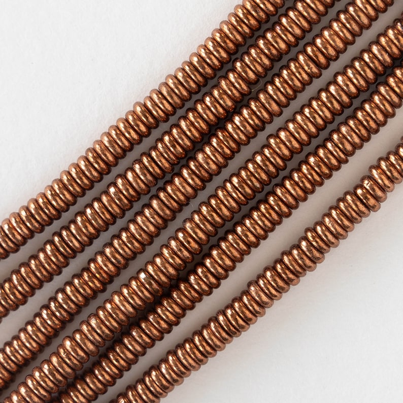 May include: A strand of copper-colored beads, each bead is a small, round coil. The beads are arranged in a diagonal line, with the top of the line extending to the right and the bottom of the line extending to the left.