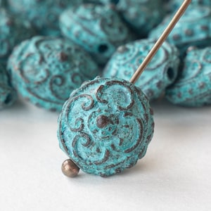 May include: Close-up of turquoise beads with an aged, textured finish. The beads have a detailed, embossed pattern and are strung on a metal needle. The beads are approximately 2.5 cm in diameter, ideal for jewelry making.
