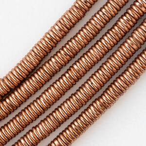Pu&ograve; includere: Primo piano di diversi fili di perline arrotolate color rame. Le perline sono uniformemente distanziate e hanno una consistenza a coste, creando un effetto a spirale. Il colore caldo e metallico delle perline contrasta con lo sfondo bianco.