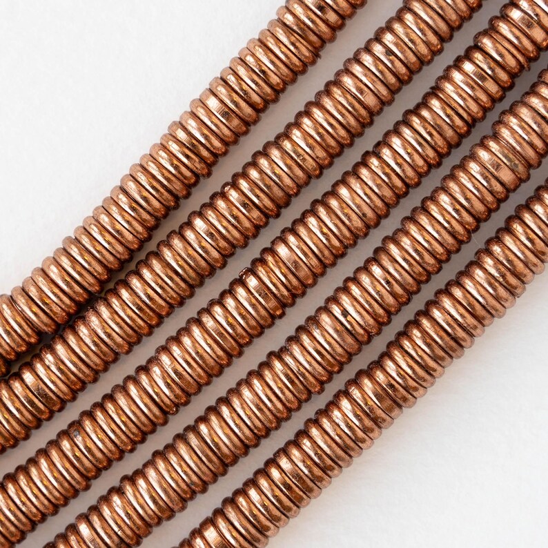 Pu&ograve; includere: Primo piano di diversi fili di perline arrotolate color rame. Le perline sono uniformemente distanziate e hanno una consistenza a coste, creando un effetto a spirale. Il colore caldo e metallico delle perline contrasta con lo sfondo bianco.