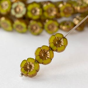 May include: Close-up of flower-shaped beads in shades of yellow and gold. The beads are strung on a silver needle, with several more beads visible in the background. The beads have a detailed, textured design, perfect for jewelry making.