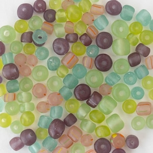 Puede incluir: Una colección de pequeñas cuentas de vidrio redondas y cilíndricas en varios tonos de púrpura translúcido, verde lima, menta y melocotón suave, con algunas cuentas que presentan finas rayas naranjas o verdes en sus superficies lisas.