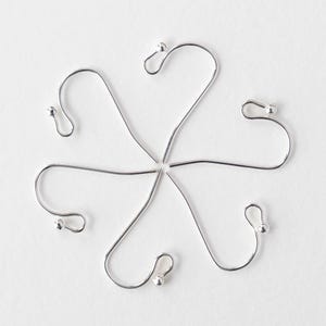 May include: Six silver-coloured ornament hooks arranged in a circular pattern. Each hook has a curved shape with a small ball at the end and a loop at the top. The hooks are on a white background.
