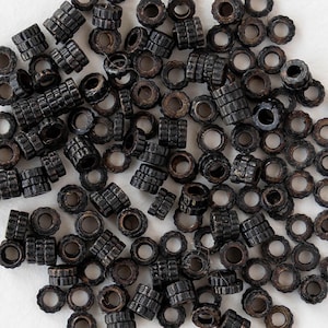 Op de afbeelding: Een verzameling kleine, donkerbruine kralen met een metallic glans. De kralen hebben verschillende vormen, waaronder cilindrische en ringvormige ontwerpen. Het oppervlak van de kralen heeft een getextureerd, geribbeld uiterlijk.