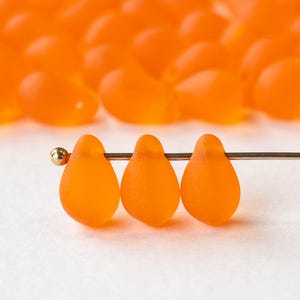 May include: Close-up of three orange teardrop-shaped beads strung on a thin wire, with a gold bead at the end. The beads have a frosted appearance. More orange beads are blurred in the background.