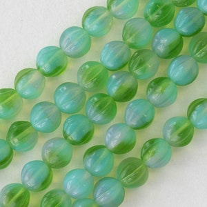 Puede incluir: Una hebra de cuentas redondas de vidrio verde y azul con un patrón estriado. Las cuentas son translúcidas y tienen un ligero brillo iridiscente.