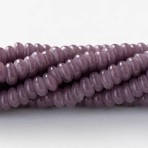 May include: A strand of purple glass beads with a slightly iridescent finish. The beads are round and have a slightly flattened shape. They are strung on a clear thread.
