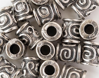 Peut inclure: Gros plan sur des perles métalliques argentées. Chaque perle cylindrique présente des motifs complexes en spirales et volutes. Les perles sont percées d'un trou central, probablement pour la création de bijoux.