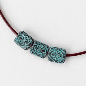 May include: A close-up of a necklace featuring three square, turquoise-colored beads with a floral design. The beads are strung on a dark red leather cord. The beads have a distressed, antique appearance.