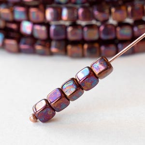 May include: Close-up of a strand of iridescent cube-shaped beads. The beads are a mix of purple, bronze, and blue hues, strung on a thin wire. The beads are approximately 0.6 cm square, with a metallic sheen. A small copper-colored bead is at the end.