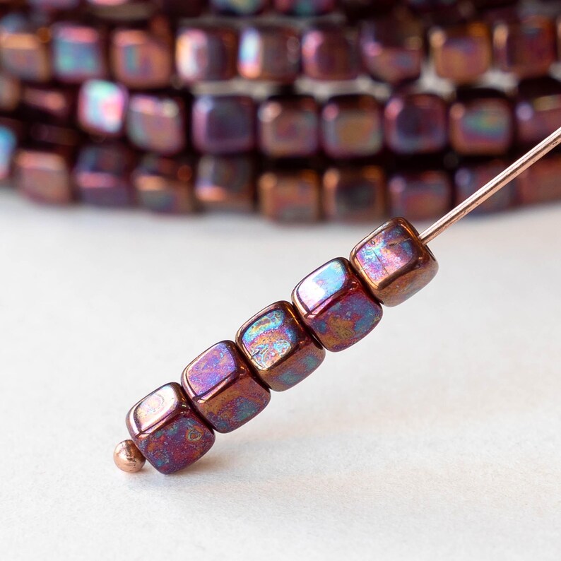 May include: Close-up of a strand of iridescent cube-shaped beads. The beads are a mix of purple, bronze, and blue hues, strung on a thin wire. The beads are approximately 0.6 cm square, with a metallic sheen. A small copper-colored bead is at the end.
