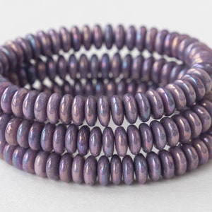 May include: Close-up of three strands of iridescent purple beads. The beads are flat, round, and stacked in rows. The beads have a metallic sheen, reflecting shades of blue and green. The background is a plain white surface.