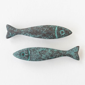 Può includere: Due sculture di pesci in bronzo con finitura verde rame. I pesci hanno una forma semplice e allungata, pinne e dettagli degli occhi. Queste sculture sono probabilmente oggetti decorativi.