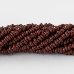 Op de afbeelding: Close-up van een streng donkerbruine houten kralen. De kralen zijn klein, rond en hebben een enigszins onregelmatige vorm. De kralen zijn aan elkaar geregen, waardoor een textuur en organisch uiterlijk ontstaat. De achtergrond is effen wit.