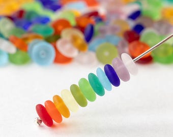 May include: A strand of colorful, flat, oval-shaped beads in a rainbow of colors, from red to purple, strung on a silver wire. A blurred background shows more beads in various colors. The beads are likely for jewelry making.