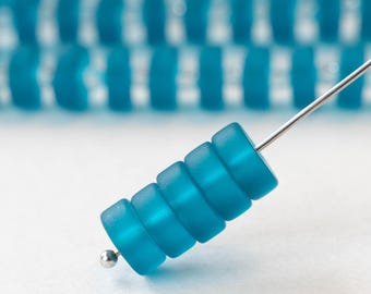 May include: A close-up of a teal-colored bead component with a silver wire. The component is made of stacked, cylindrical beads. The background shows more of the same components, out of focus.