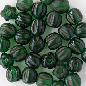 Pode incluir: Uma coleção de contas de vidro verde escuro com listras marrons e várias formas, incluindo designs redondos e em forma de flor. As contas estão espalhadas em uma superfície branca, mostrando sua textura brilhante e padrões intrincados. Ideal para a criação de joias.