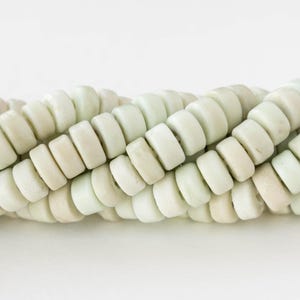 May include: Close-up of a strand of pale green and off-white beads. The beads are cylindrical with rounded edges, strung together in a twisted pattern. The beads have a matte finish and a natural, organic appearance.