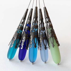 May include: An array of five glass pendants in varying shades of blue and green. Each pendant has a frosted, teardrop-shaped glass body and is topped with an ornate, antique silver-colored metal cap. The pendants are arranged in a fan shape.