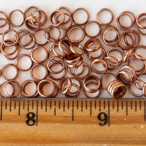 Pu&ograve; includere: Una collezione di piccoli anelli metallici color rame sparsi su una superficie. Gli anelli sono circolari con un centro cavo. Un righello di legno con segni in centimetri &egrave; visibile in basso.