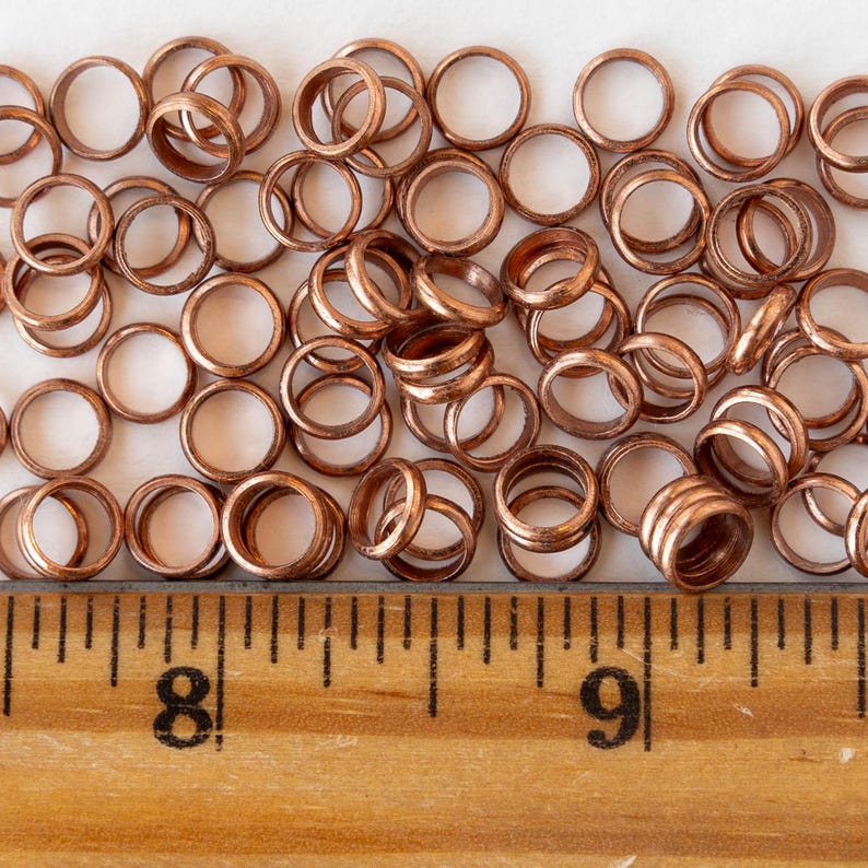 Pu&ograve; includere: Una collezione di piccoli anelli metallici color rame sparsi su una superficie. Gli anelli sono circolari con un centro cavo. Un righello di legno con segni in centimetri &egrave; visibile in basso.