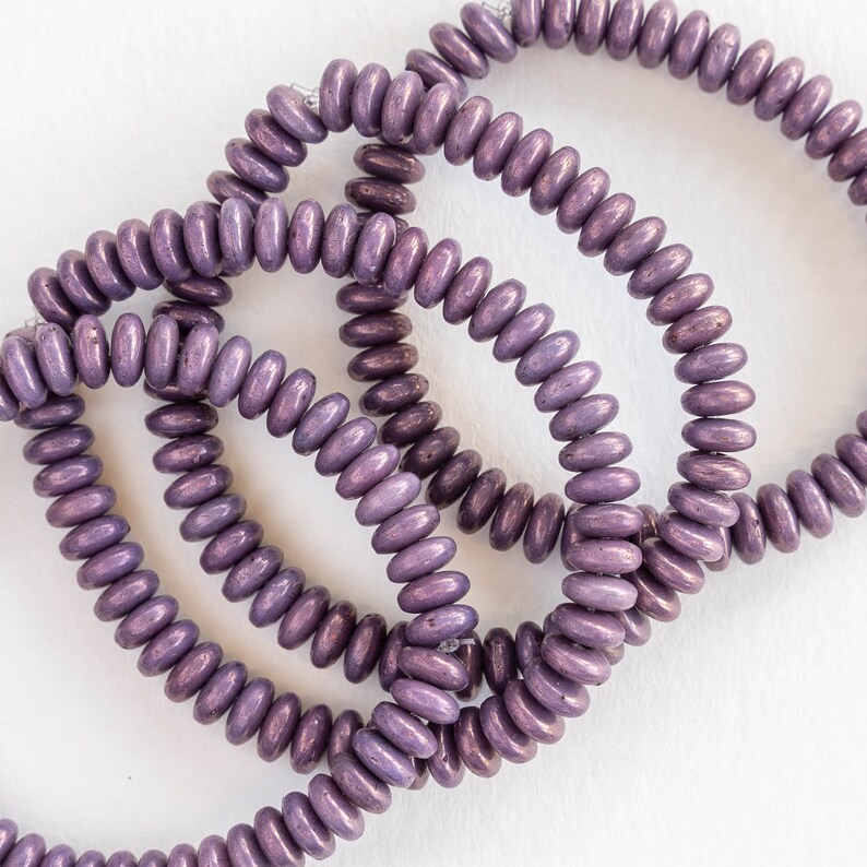 May include: Close-up of several strands of purple wooden beads. The beads are oval-shaped and strung together, creating a circular pattern. The beads have a metallic sheen, and the color varies slightly, with some appearing darker than others.