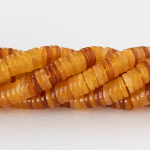 May include: Close-up of a strand of amber gemstone beads. The disc-shaped beads have light and dark amber tones. The beads are strung together, creating a textured effect. The background is plain white.