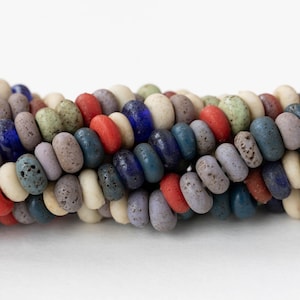 May include: A close-up of a strand of colorful, oval-shaped beads. The beads are in various colors, including red, blue, green, and cream. The beads have a textured, matte finish and are strung together.
