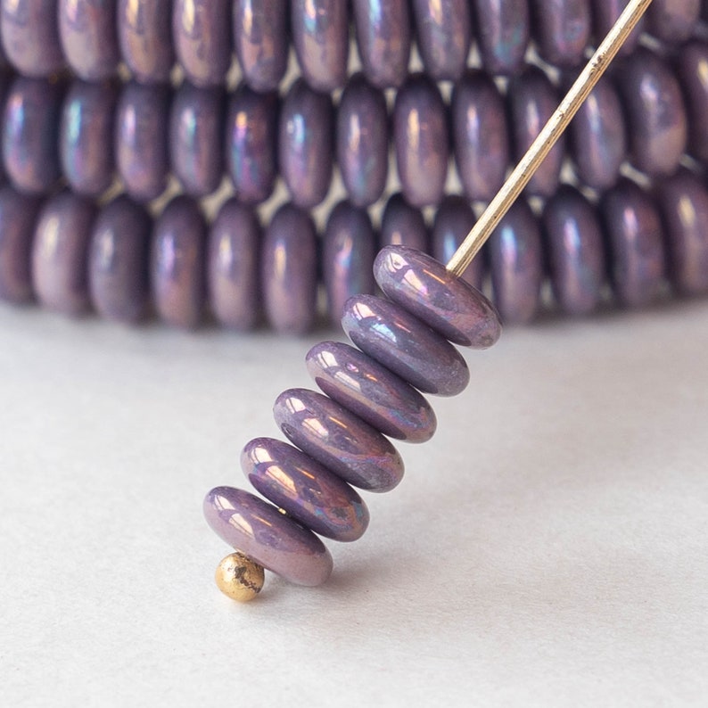 May include: Close-up of iridescent purple beads on a gold pin, with a blurred background of similar beads. The beads have a flat, oval shape and a shiny, reflective surface. The beads are likely for jewelry making.