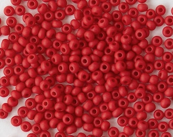May include: A large collection of small, round, matte red seed beads scattered across the frame. Each bead features a central hole for threading, displaying a consistent opaque red colour and a smooth, uniform texture throughout the entire group.