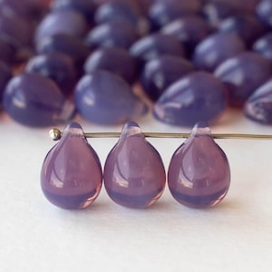 Puede incluir: Tres cuentas en forma de lágrima, en tonos morados, ensartadas en un alambre, con un fondo borroso de cuentas similares. Las cuentas tienen una cualidad translúcida, reflejando la luz. Adecuado para la fabricación de joyas.