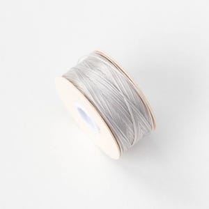 Könnte beinhalten: Eine Spule grauer Schnur, eng um einen Pappkern gewickelt. Die Schnur scheint aus einem glatten, möglicherweise gewachsten Material zu bestehen. Die Spule ist vor einem schlichten weißen Hintergrund platziert, der das Produkt hervorhebt.