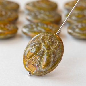 May include: Close-up of a golden-green oval bead with a floral design. The bead is strung on a silver needle, with several similar beads in the background. The beads have an iridescent finish.