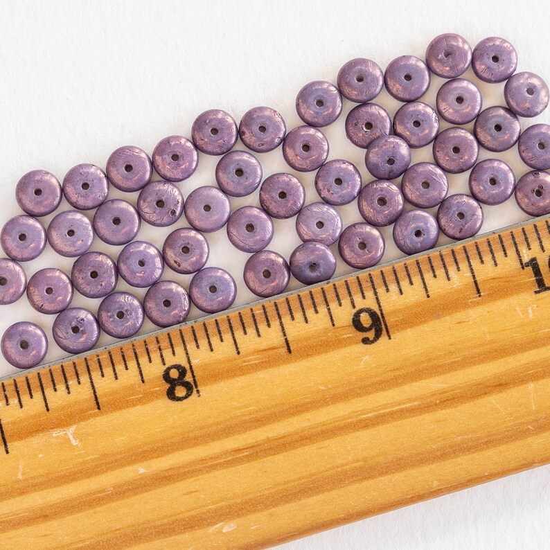 May include: Close-up of a collection of small, flat, round beads in a muted purple hue. Each bead has a central hole and a metallic sheen. A wooden ruler with inch markings is in the foreground, providing scale.