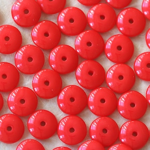 Puede incluir: Una gran cantidad de cuentas redondas de color rojo brillante con un agujero en el centro. Las cuentas están dispuestas en filas sobre una superficie marrón claro.