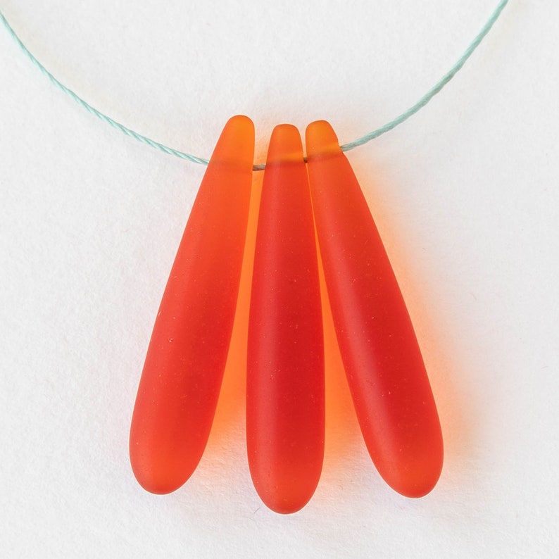 Puede incluir: Tres cuentas de vidrio en forma de l&aacute;grima de color naranja brillante. Las cuentas est&aacute;n enhebradas en un cord&oacute;n blanco delgado.