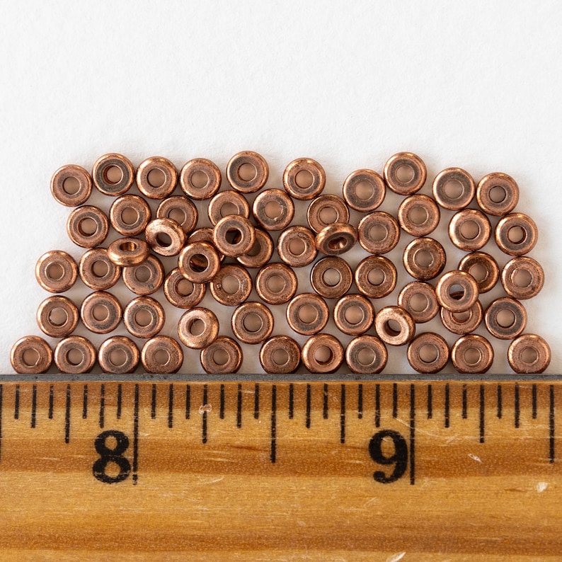 May include: A pile of copper-colored metal beads, arranged in a loose, uneven pattern. The beads are small and round, with a hole in the center. The beads are arranged on a white surface, with a wooden ruler in the foreground. The ruler is marked with inches, and the numbers 8 and 9 are visible.