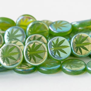 May include: A collection of green glass pebbles with a cannabis leaf design. The pebbles have a clear, iridescent finish and are scattered across a white surface. The leaves are a dark green color.