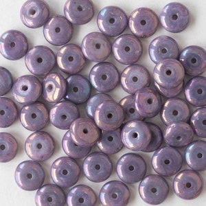 May include: A collection of iridescent lavender-colored glass beads. Each bead is round with a central hole, and the surface reflects a subtle rainbow sheen. The beads are scattered on a white surface, ideal for jewelry making or craft projects.