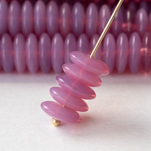 Puede incluir: Una pila de cinco cuentas de vidrio rosa, cada una con un tono de rosa ligeramente diferente, están enhebradas en un alambre dorado. Las cuentas son de forma ovalada y tienen un acabado liso y brillante.