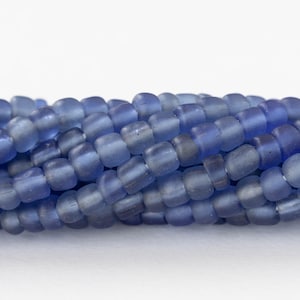 Puede incluir: Primer plano de un hilo de cuentas de vidrio azules. Las cuentas son cilíndricas con bordes redondeados y un acabado mate. El color es un azul apagado, con variaciones de tono. Las cuentas están ensartadas, creando una superficie texturizada.