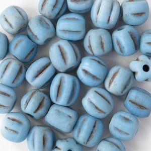 May include: A collection of light blue, barrel-shaped beads with brown, grooved accents. The beads are scattered across a white surface. One bead is flower-shaped. These beads are suitable for jewelry making and crafts.