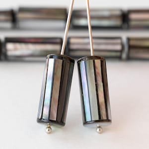 Op de afbeelding: Twee cilindrische kralen met verticale inlays van iriserend zwart parelmoer. Elke kraal is gemonteerd op een dunne zilverkleurige metalen pin met een kleine bolvormige kraal aan de basis, met een donker, reflecterend oppervlak.