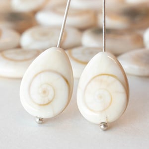 May include: Two white teardrop-shaped shell beads with a spiral pattern in the center. The beads are strung on silver wires.