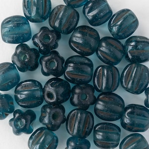 May include: An assortment of teal-colored glass beads. The beads vary in shape, including round, flower-shaped, and ribbed designs. The beads are scattered across a white surface, showcasing their translucent quality.